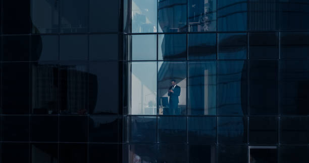 istock Aerial View Shot: From Outside into Office Building with Businessman Using Mobile Phone and Standing Next to the Office Window. Beautiful Shot of The Financial Business District Skyscrapers. 1069160390