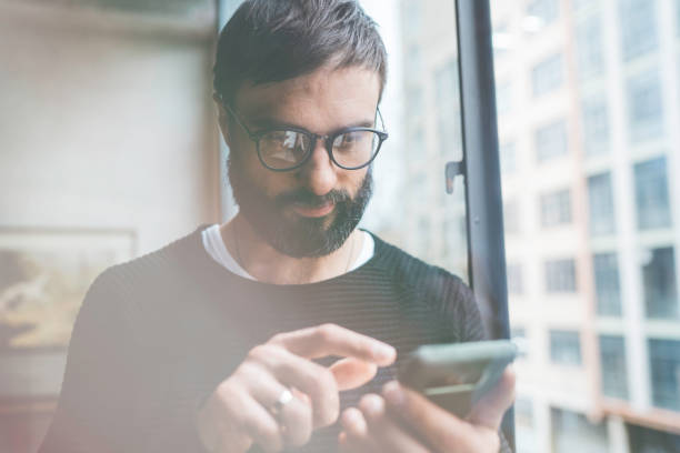 istock Handsome bearded man wearing eye glasses using mobile smartphone for surfing web internet. Man using gadget at modern loft studio. Horizontal. Blurred background. 1069175360