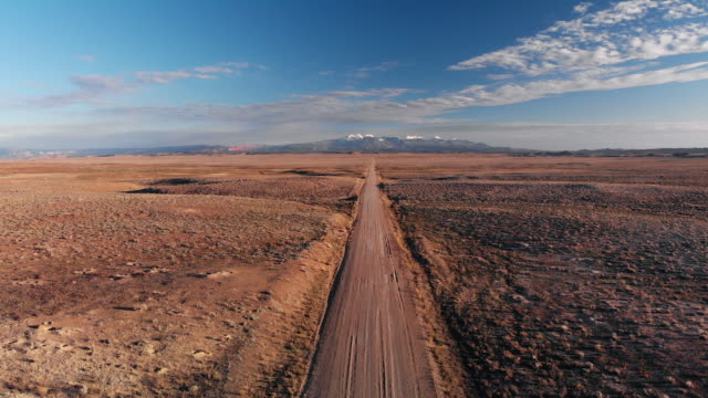 Ascending Aerial Drone Shot of a Vanishing Point Dirt Road with Mountains in the Background Outside of Moab, Utah with Desert Plains on Either Side Underneath a Blue Sky at Sunset/Sunrise
