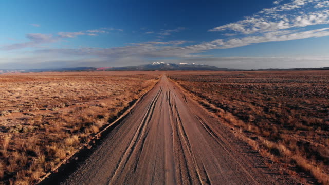 Moving Forward Aerial Drone Shot of a Vanishing Point Dirt Road with Mountains in the Background Outside of Moab, Utah with Desert Plains on Either Side Underneath a Blue Sky at Sunset/Sunrise