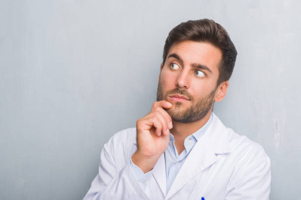 istock Handsome young professional man over grey grunge wall wearing white coat with hand on chin thinking about question, pensive expression. Smiling with thoughtful face. Doubt concept. 1069185080