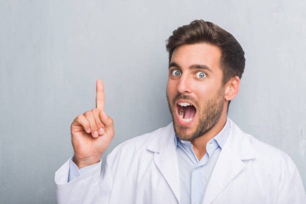 istock Handsome young professional man over grey grunge wall wearing white coat pointing finger up with successful idea. Exited and happy. Number one. 1069185082