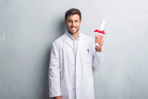 Photo of Handsome young doctor man over grey grunge wall holding diploma with a happy face standing and smiling with a confident smile showing teeth