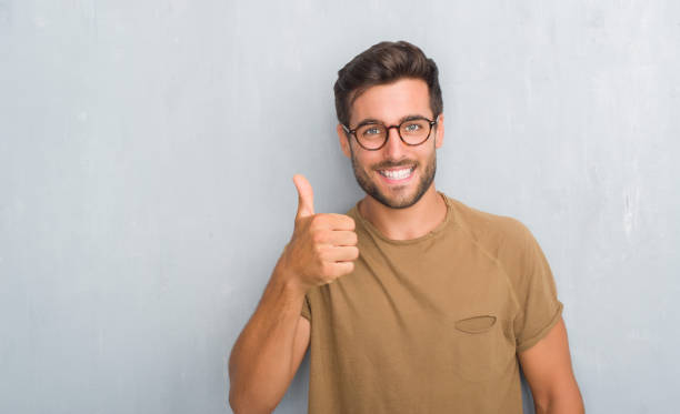 beau jeune homme sur mur gris grunge lunettes faisant le geste avec la main pouce en l’air heureux. approuvant l’expression en regardant la caméra avec la liste des succès. - doigts en forme de o photos et images de collection