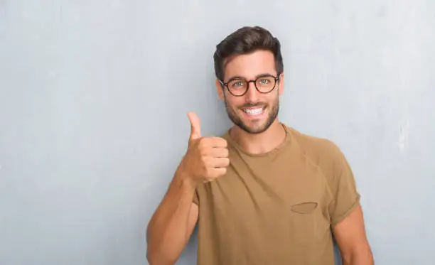 Photo of Handsome young man over grey grunge wall wearing glasses doing happy thumbs up gesture with hand. Approving expression looking at the camera with showing success.