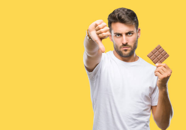 istock Young handsome man eating chocolate bar over isolated background with angry face, negative sign showing dislike with thumbs down, rejection concept 1069188502