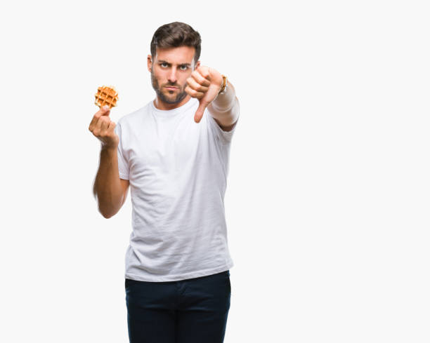 istock Young handsome man eating a sweet waffle over isolated background with angry face, negative sign showing dislike with thumbs down, rejection concept 1069189406