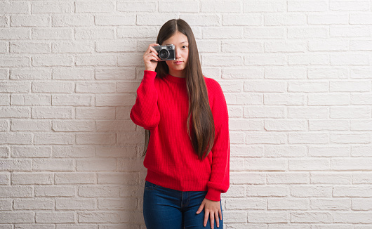 Young Chinese woman over brick wall holding vintage camera with a confident expression on smart face thinking serious