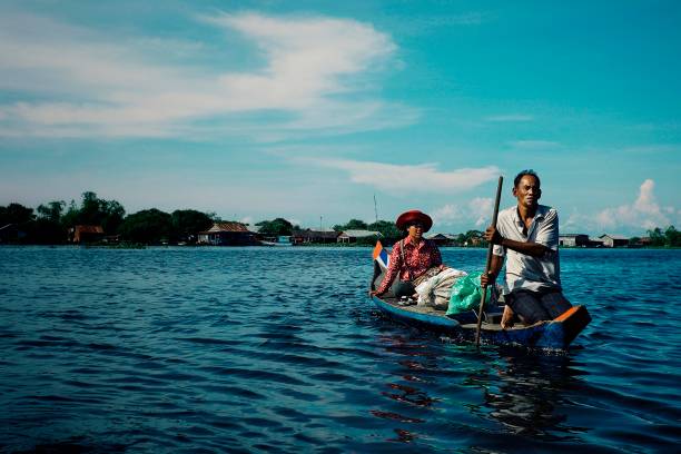 famiglia locale attraversando il lago sulla sua canoa di fronte all'insediamento galleggiante palafitte - flood people asia cambodia foto e immagini stock
