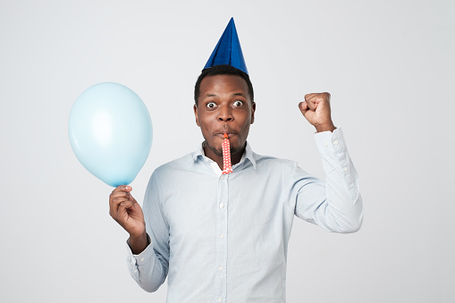 Handsome cheerful young african man having fun on party wearing blue shirt and holiday hat, blowing party horn. People, joy, birthday and celebration concept