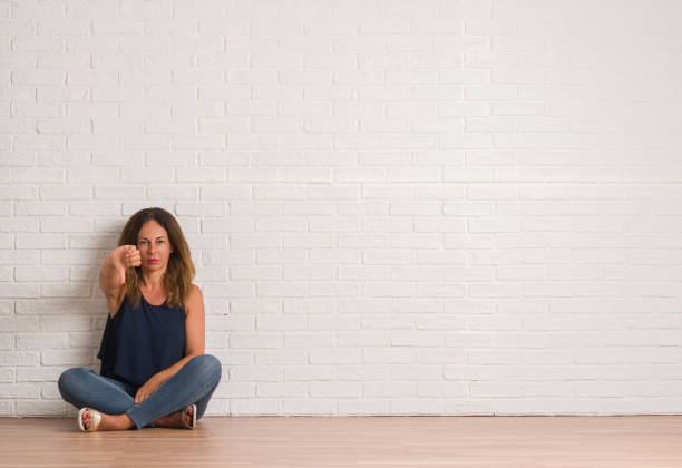 istock Middle age hispanic woman sitting on the floor over white brick wall looking unhappy and angry showing rejection and negative with thumbs down gesture. Bad expression. 1069269014
