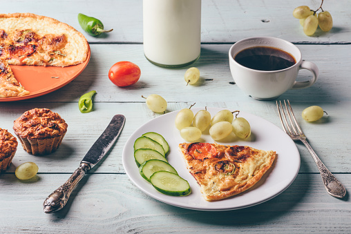 Breakfast frittata with chorizo, tomatoes and chili peppers on plate with cup of coffee, grapes and muffins over light wooden background. Healthy eating concept.
