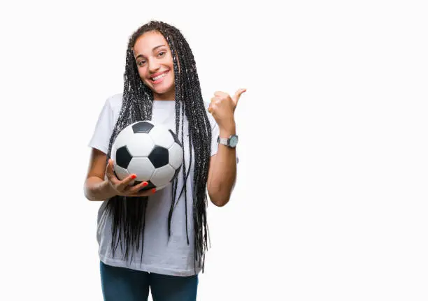 Photo of Young braided hair african american girl holding soccer ball over isolated background pointing and showing with thumb up to the side with happy face smiling