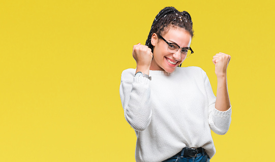 Young braided hair african american girl wearing glasses and sweater over isolated background very happy and excited doing winner gesture with arms raised, smiling and screaming for success. Celebration concept.