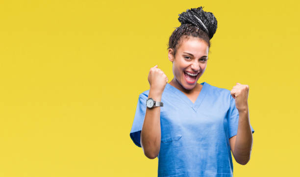 młoda pleciona włosy african american dziewczyna profesjonalna pielęgniarka na odizolowanym tle bardzo szczęśliwy i podekscytowany robi gest zwycięzcy z rękami podniesionymi, uśmiechnięty i krzycząc o sukcesie. koncepcja celebracji. - cheerful happiness surgeon success zdjęcia i obrazy z banku zdjęć
