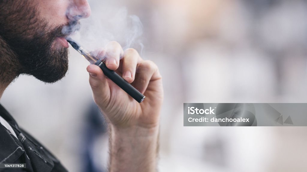 Young male worker smoking electronic cigarette Smoking electronic cigarette. Shallow DOF. Developed from RAW; retouched with special care and attention; Small amount of grain added for best final impression. 16 bit Adobe RGB color profile. Electronic Cigarette Stock Photo