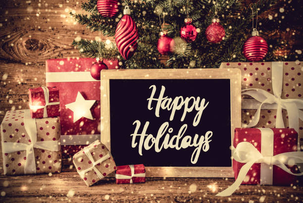 Chalkboard With English Calligraphy Happy Holidays. Christmas Tree With Christmas Tree Ball Ornament. Presents And Gifts On Rustic Wooden Background With Snowflakes