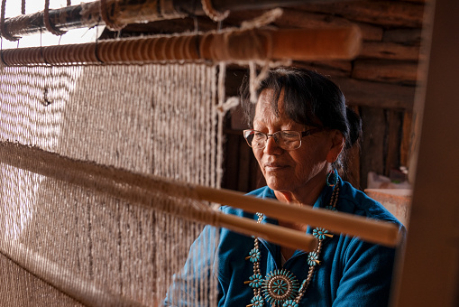Traditional Navajo elderly woman working on a blanket loom in an authentic hogan