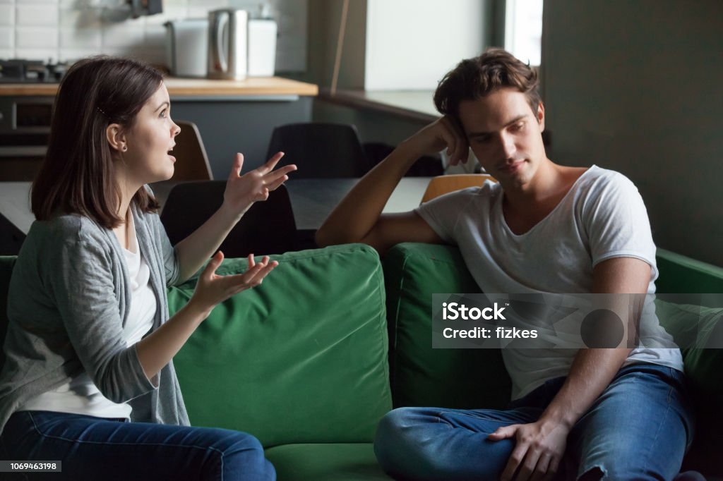 Young couple talking about their relations at home Young attractive couple sitting on couch at home and sorting out their relations. Girlfriend talking too much, drama queen nagging disinterested boyfriend feels angry. Troubles and break up concept Couple - Relationship Stock Photo