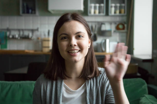 Attractive girl sitting on couch looking at camera Head shot portrait of cheerful young woman sitting on sofa at home. Girl in casual clothes have video call using computer, wave her hand greeting or say goodbye to friend and smiling looking at camera divergent movie review stock pictures, royalty-free photos & images
