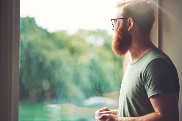 Where will I be a year from now? Shot of a young man having coffee and looking out of a window at home looking at view stock pictures, royalty-free photos & images