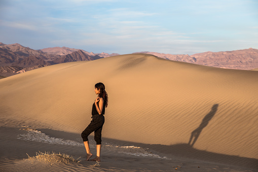 Beautiful mixed race young woman standing in  golden sand dunes before sunset facing to the side. Large shadow cast on sand dune. Death Valley Mesquite Flat sand dunes