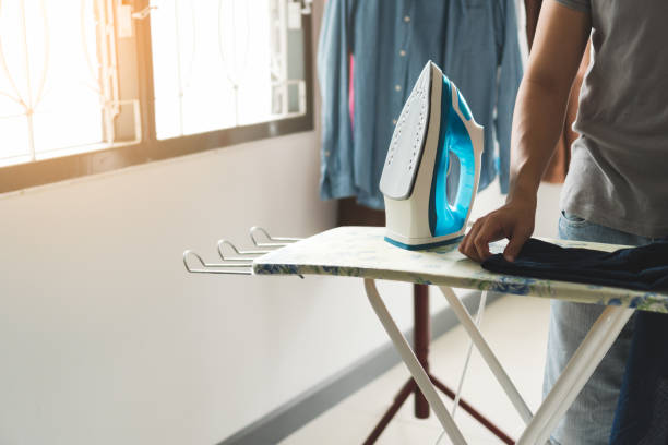 Young asian man ironing jean clothes on iron board with steaming blue iron at home. Young asian man wearing gray t-shirt ironing jean clothes on iron board with steaming blue iron at home. Man in role of a woman duty. Housework and household concept. laundry husband housework men stock pictures, royalty-free photos & images