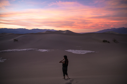 Beautiful sunset with young petite woman walking down sand dune footprints leading away in Death Valley. Large dunes in the distance. One woman only. Hand in hair