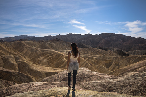 Beautiful woman with white shirt wearing sunglasses standing looking at unique hill patterns formations in Death Valley Zabriskie Point. Bright with mountains in background and blue clear sky.