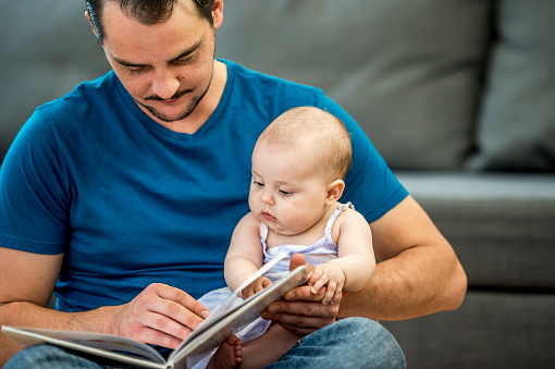 A man has his baby daughter in his lap as they turn the pages of a storybook he is reading from.