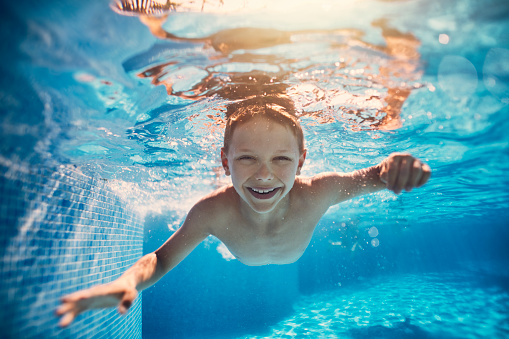 Portrait of smiling little boy swimming underwater in the pool towards the camera. Sunny summer day.\nNikon D850