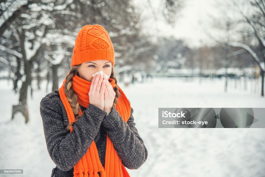 Cold A young woman with braided long blond hair is outdoors in a park during the winter. There is snow and trees in the background. She is wearing winter clothes, an orange hat and scarf, and is blowing her nose with a handkerchief / hanky / tissue. She is looking away from the camera. With copy space. Winter Stock Photo