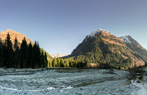 rocky mountain landscape in early winter with snow-capped peaks and colorful forest and a mountain stream in the foreground with ice and rime on the rock and grass