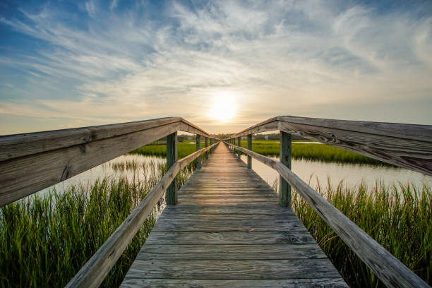 sunset over coastal waters with a very long wooden boardwalk coastal waters with a very long wooden boardwalk pier in the center during a colorful summer sunset under an expressive sky with reflections in the water and marsh grass in the foreground tidal inlet stock pictures, royalty-free photos & images