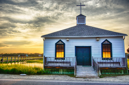 small white wooden chapel on the water on the coast during a colorful summer sunset with illuminated golden windows and a setting sun under a blue sky with soft clouds