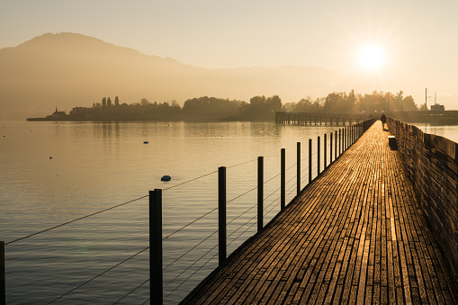 long wooden pier and boardwalk over Lake Zurich near Rapperswil in golden evening light with silhouette of pedestrians and people walking and mountains in the background