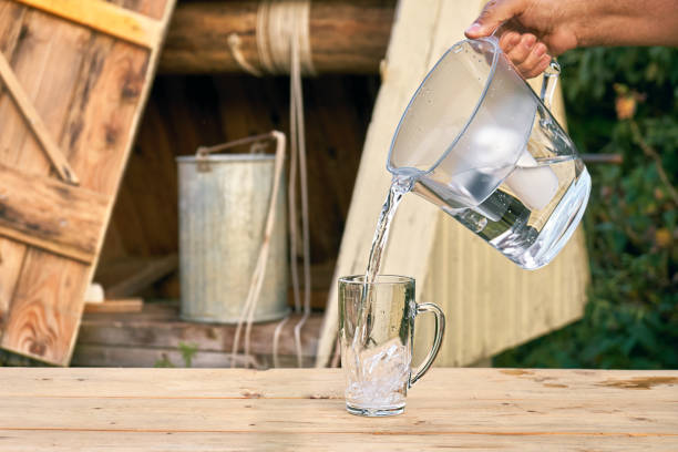 Pouring a clean filtered water from a water filter jug to a glass cup in front of wooden draw well stock photo