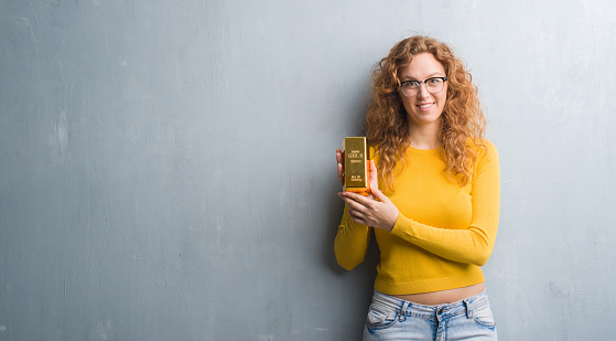 Young redhead woman over grey grunge wall holding gold ingot with a happy face standing and smiling with a confident smile showing teeth