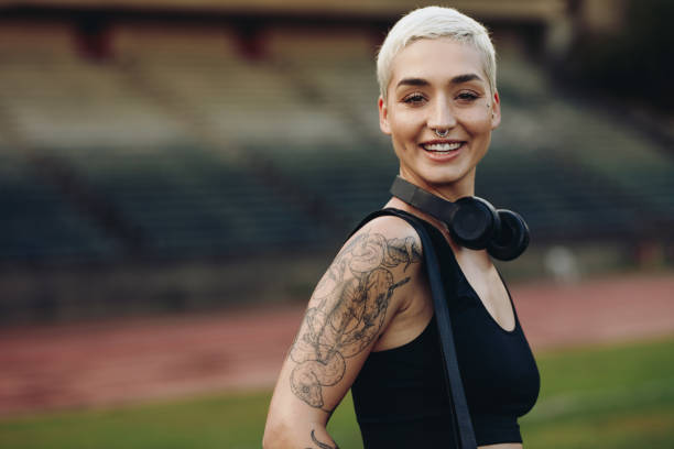 Cheerful fitness woman inside an athletic stadium with her bag and wireless headphones. Side view of a female athlete standing in a track and field stadium.