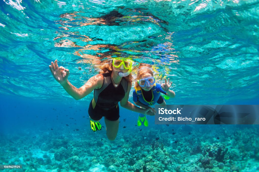Mother, kid in snorkeling mask dive underwater with tropical fishes Happy family - mother, kid in snorkeling mask dive underwater with tropical fishes in coral reef sea pool. Show by hands divers sign OK. Travel lifestyle, beach adventure on summer holiday with child. Snorkeling Stock Photo