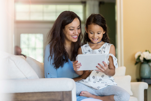 Smiling mid adult mom and her adorable elementary age daughter smile as they watch something on a digital tablet.