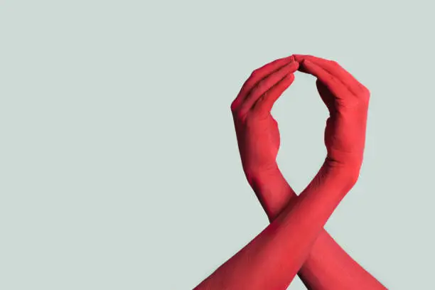 closeup of the arms of two men painted red forming a red awareness ribbon for the fight against AIDS, against an off-white background with some blank space on the left