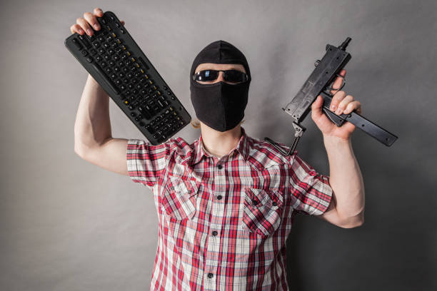 Crazy hacker man. Unrecognizable guy wearing black balaclava holding gun and keyboard. Hate speech on the internet.