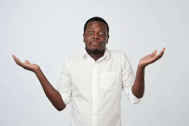 Young african man in white shirt being at a loss, showing helpless gesture with arm and hands, as if he does not know what to do. Why did I fail my job.