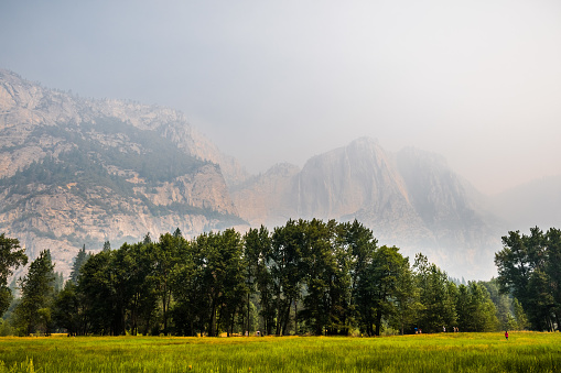 Landscape in Yosemite Valley; green meadow in the foreground; rock walls and Yosemite Upper Falls visible in the background through smoke from Ferguson Fire; Yosemite National Park, California