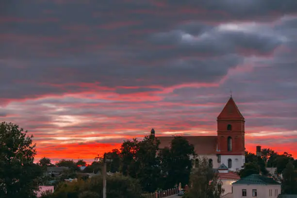 Photo of Mir, Belarus. Saint Nicolas Roman Catholic Church In Backlight Of Amazing Sunset Over Landscape Of Village Houses. Famous Landmark