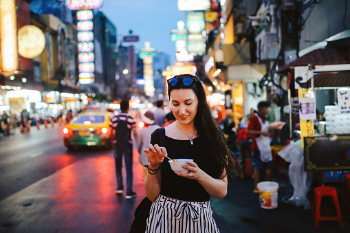 Portrait of a young brunette woman, walking on the streets of Bangkok Chinatown district. She is wearing casual street style clothing, trying out Thai food on the go. Delicious deep fried shrimp tempura dipped in hot sauce.