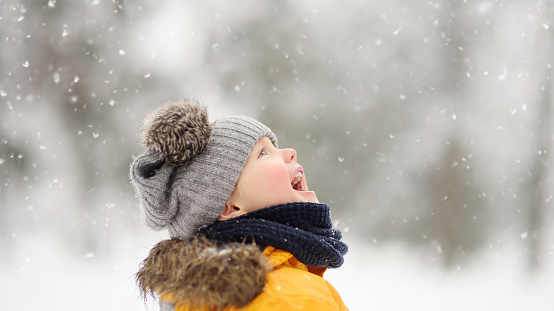 Cute little boy in yellow winter clothes walks in during a snowfall. Outdoors winter activities for kids.