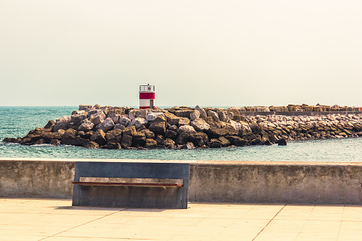 Small Lighthouse with red and white stripes in Marina de Oeiras. View with a beautiful metallic street bench and some harbour rocks called breakwaters.