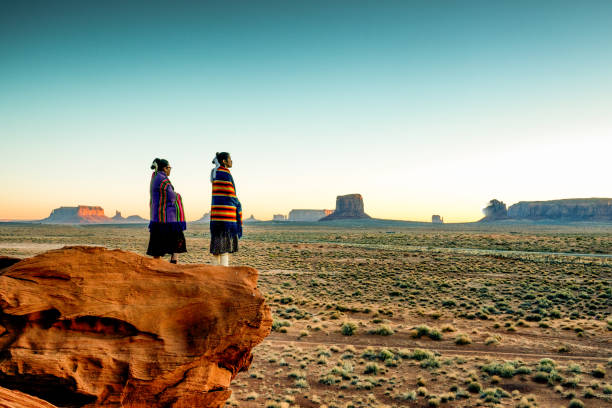 Two Traditional Navajo Native American Sisters In Monument Valley Tribal Park on a Rocky Butte Enjoying a Sunrise or Sunset Pretty Navajo girls wrapped in handwoven traditional blankets enjoying a grand sunrise or sunset in Monument Valley indigenous peoples of the americas stock pictures, royalty-free photos & images